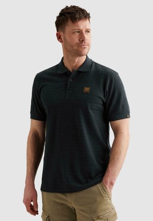 Uomo con polo nera a maniche corte con un piccolo logo sul petto e pantaloncini cargo kaki, in piedi con una mano in tasca.