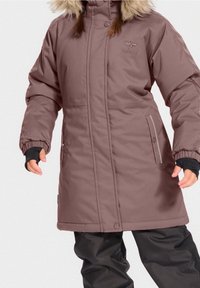 Manteau d'hiver long, marron, avec une capuche en fourrure, des poignets élastiques et des poches latérales. Présente une texture lisse et un détail de logo sur le devant.