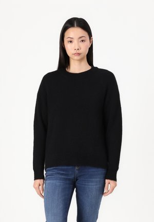 Schwarzer Pullover mit Rundhalsausschnitt, leicht oversized Passform und strukturiertem Stoff. Model kombiniert mit blauen Jeans, vor einem weißen Hintergrund.