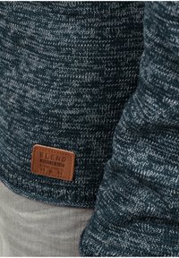 Tmavomodrý pletený sveter s kombináciou svetlých a tmavých nití. Má hnedú koženú štítok s nápisom „BLEND“ a dátumom založenia. Textúrovaná látka.