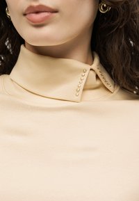 Gros plan d'une femme portant un col roulé beige avec un col pointu orné de petits clous dorés et des boucles d'oreilles créoles en or.