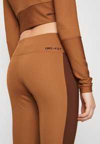 Hnědé legíny Dri-Fit s texturovaným žebrovaným povrchem; mají kontrastní panely ve tmavě hnědé barvě a logo na pásku.