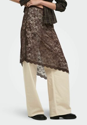 Personne portant un pantalon beige à jambes larges sous une jupe en dentelle marron transparente, associée à des chaussures noires et un haut à carreaux.