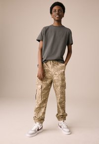 T-shirt grigia in cotone, pantaloni cargo kaki con motivo mimetico e sneakers bianche con dettagli marroni. Vestibilità casual e polsini elastici.