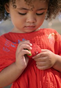 Rotes Baumwollkleid mit strukturiertem Stoff und bestickten Details; Kind hält ein weiches, plüschiges Erdbeer-Spielzeug in den Händen.