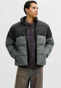 Vadderad jacka i svart och mörkgrönt. Har hög krage, dragkedja framtill och quiltad design. Bärs över en mörk tröja och slitna jeans.