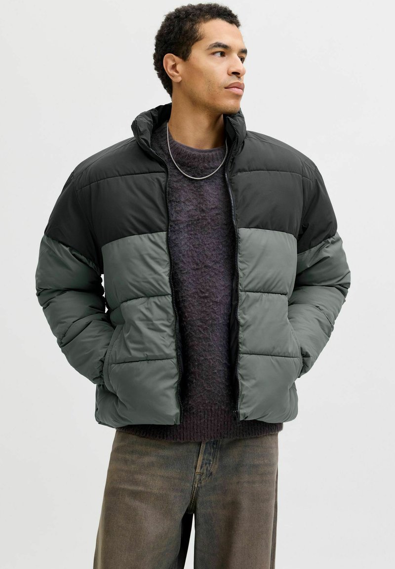 Vadderad jacka i svart och mörkgrönt. Har hög krage, dragkedja framtill och quiltad design. Bärs över en mörk tröja och slitna jeans.