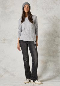 Grijze geribde trui met lange mouwen, gecombineerd met een donkere spijkerbroek met een koord. Model draagt een bijpassende grijze beanie en witte sneakers.