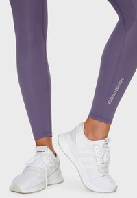 Ljubičaste leggings koje ne proziru sa uskim fitom i ravnim šavovima, u kombinaciji s bijelim sportskim cipelama koje imaju tri pruge i teksturiranu gornjište.