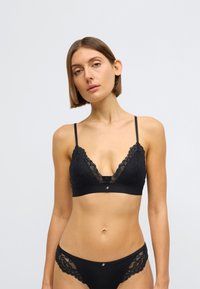 Crna čipkasta bralet s podesivim naramenicama, ukrašenim nazubljenim rubovima, prozirnim panelom i metalnim logotipom. Usklađene crne čipkaste gaćice.