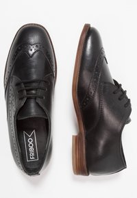 Černé kožené boty s brogue vzorem, které mají perforované detaily a ploché dřevěné podpatky. Jsou vidět šněrování a hladký povrch.