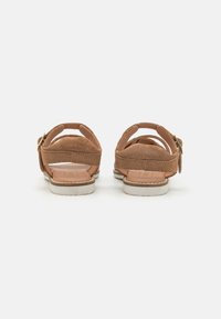 Brune semskede sandaler med bagrem og gyldne hardware-detaljer, med en flad hvid sål og tekstureret fodseng.
