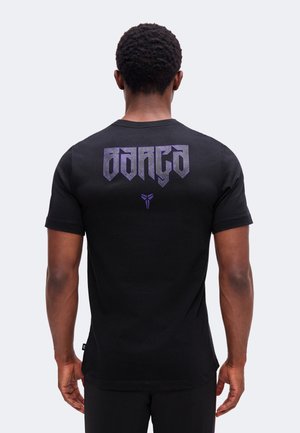 Homme aux cheveux courts et bouclés vu de dos, portant un t-shirt noir avec un texte stylisé violet et un logo sur le haut du dos.