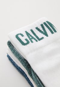 Calcetines Calvin Klein apilados, con una base blanca y texto verde y acentos azul verdoso. Textura suave con detalle de ribete en la parte superior.