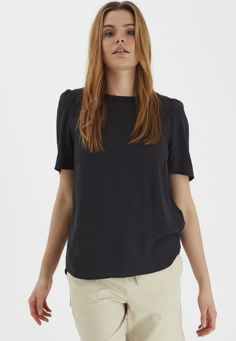Top corto nero a maniche corte con scollatura quadrata, texture liscia e vestibilità rilassata, abbinato a pantaloni chiari. Cuciture di un colore leggero.
