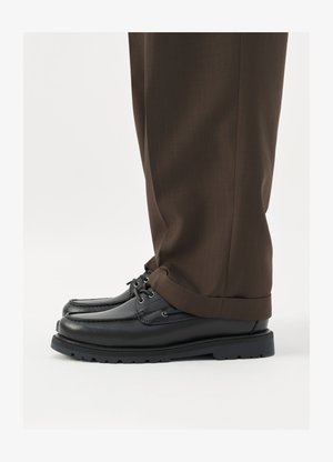 Sapatos de pele preta com bico arredondado, atacadores e sola de borracha grossa, combinados com calças sociais castanhas.