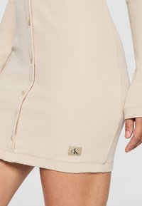 Beige ribbad klänning med knäppning framtill, långa ärmar och en märkesetikett i fållen. Slät textur, midjehöjd.