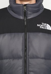 Sort og grå isoleret jakke med høj krave, lynlåslukning og broderet logo på brystet. Materialet virker glat med et polstret design.