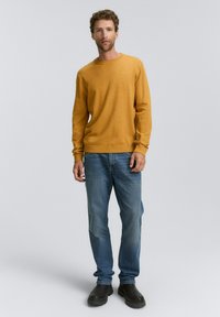 Suéter mostaza, cuello redondo, con puños acanalados, combinado con jeans azules y zapatos negros de slip-on, de pie contra un fondo gris claro.