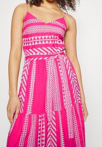 Robe rose avec des bretelles spaghetti fines, présentant un motif géométrique en blanc et rose, taille cintrée et jupe fluide avec du tissu froncé.