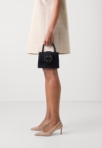 Femme portant une jupe à carreaux beiges et des escarpins nude, tenant un petit sac à main noir avec une boucle en argent dans sa main droite.