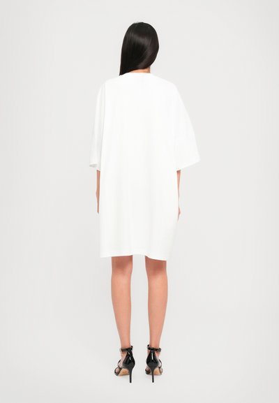 Weißes, übergroßes T-Shirt-Kleid, mit kurzen Ärmeln, aus Baumwolle, mit geradem Schnitt; getragen mit schwarzen High Heels mit glatter Textur.