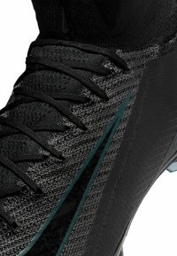 Scarpa sportiva nera caratterizzata da una tomaia in maglia testurizzata, spesse stringhe nere e un design elegante con un accento verde a contrasto.