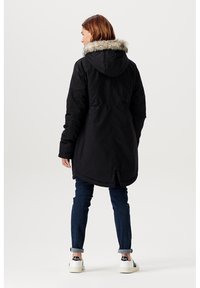 Černá parka s kapucí lemovanou kožešinou, s delším zadním lemem, nošená s ohrnutými džínami a bílými teniskami.