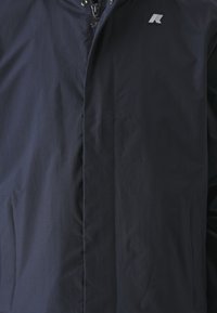 Giacca isolata blu navy con zip frontale, tasche laterali e patch del logo. Tessuto liscio con un leggero lucido.