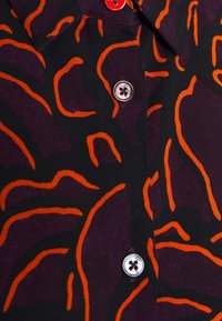 Tissu noir avec un motif abstrait violet et orange, boutonné avec un bouton argenté et un bouton accent rouge au col.
