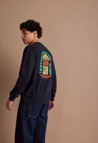 Marineblauwe sweatshirt met kleurrijke grafische tekst en zonontwerp op de achterkant. Comfortabele stof, geribbelde boorden en zoom, gecombineerd met losse denim jeans.