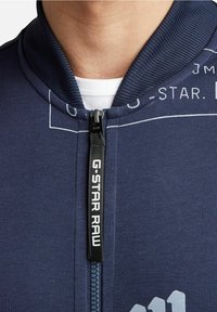 Chaqueta de cremallera navy con cuello acanalado, acentos de logo en blanco y una textura de tela suave. El tirador de la cremallera muestra "G-STAR RAW".