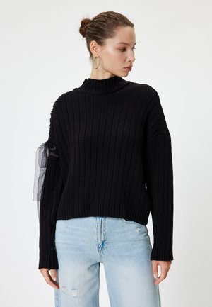 Zwarte geribde trui met een hoge nek en lange mouwen, voorzien van een doorschijnende zwarte tuleaccent op de linkermouw, gecombineerd met lichtblauwe jeans.