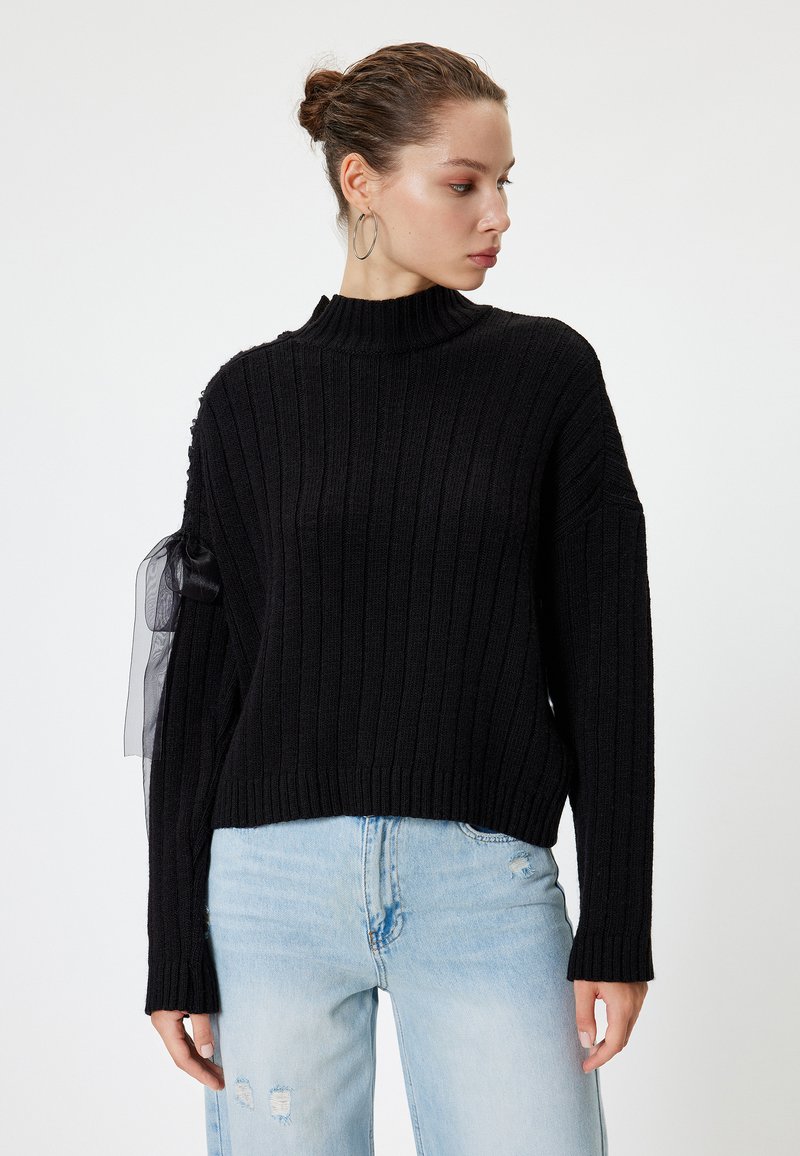 Zwarte geribde trui met een hoge nek en lange mouwen, voorzien van een doorschijnende zwarte tuleaccent op de linkermouw, gecombineerd met lichtblauwe jeans.