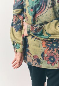 Kleurvol, gestreept shirt in groen, rood en blauw met bloemenpatroon, voorzien van lange mouwen en knoopmanschetten. Draagt met zwarte shorts.