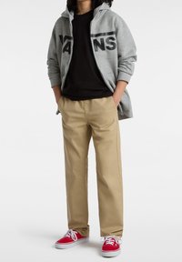 Šedá mikina na zip s logem "VANS", černé tričko, béžové kalhoty a červené tenisky s bílými tkaničkami. Hladký materiál a volný střih.