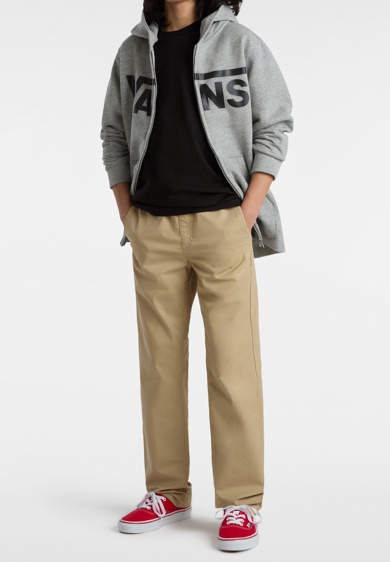 Šedá mikina na zip s logem "VANS", černé tričko, béžové kalhoty a červené tenisky s bílými tkaničkami. Hladký materiál a volný střih.