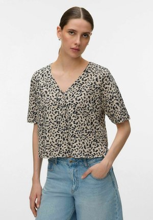 Kobieta z zaczesanymi do tyłu włosami, ubrana w beżową bluzkę z dekoltem w kształcie litery V z czarnymi abstrakcyjnymi plamami oraz jasnoniebieskie, dżinsy z wysokim stanem, ręka w kieszeni.