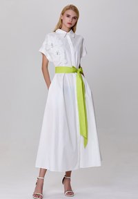Robe blanche en coton avec des manches courtes, détails en appliqué floral et une ceinture verte vibrante nouée à la taille. Comprend des poches latérales.