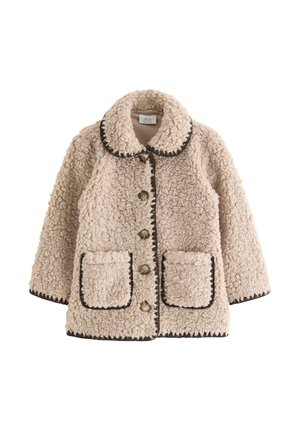 Jeune fille souriante, portant une veste sherpa beige avec des boutons, un t-shirt rose avec un dessin animé, un pantalon à carreaux noir et blanc et des baskets bordeaux.