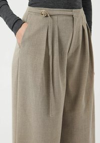 Pantaloni beige chiaro sartoriali con vita alta, pieghe e dettaglio circolare in metallo dorato. Il tessuto appare strutturato e morbido, con tasche laterali.