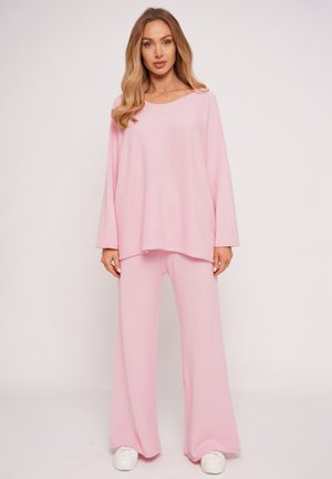 Kobieta stojąca w luźnym, jasnoróżowym topie z długim rękawem i szerokich spodniach z białymi trampkami na białym tle.