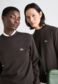 To personer har på seg mørkebrune collegegensere med en grønn Lacoste-logo. En av dem bærer en grønn håndveske med skulderstropp.