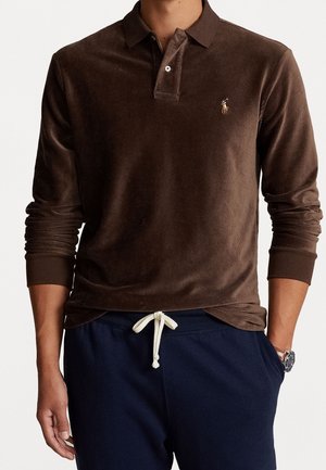 Homme portant un polo à manches longues en velours côtelé marron avec un pantalon à cordon de serrage bleu marine et une montre au poignet gauche.