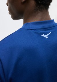 Άτομο που φαίνεται από πίσω φοράει μπλε πουκάμισο με ένα μικρό λευκό λογότυπο κοντά στον λαιμό στο πάνω μέρος της πλάτης.