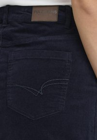 Temno modri džins hlače z teksturirano površino. Imajo en zadnji žep s kontrastnim šivom in blagovno znamko iz usnja na pasu.