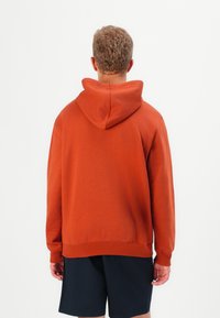 Hoodie rust narančaste boje s kapuljačom koja se može prilagoditi, s rebrastim manžetama i rubom. Jednostavan dizajn, napravljen od mekog materijala, nosi se preko tamnih šortseva.