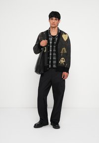 Cárdigan de punto negro con diseños coloridos, superpuesto a una camisa de cuadros, combinado con pantalones cargo negros holgados y zapatos negros. Bolsa negra.