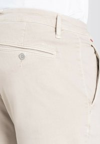 Beige cotton trousers featuring a back pocket with a zipper and a logo embossed on the waistband. The fabric has a smooth texture.