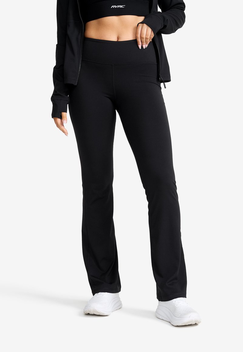 Svarta träningsbyxor med hög midja, tillverkade av stretchigt tyg, med en slät yta och utställd ben-design, i kombination med vita sneakers.